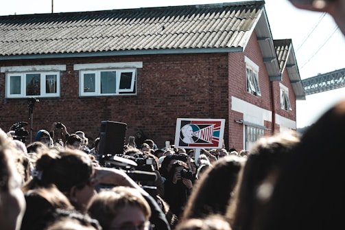 A large crowd of people gathered outdoors, many holding cameras or their phones aloft, appears to be at some sort of public event or protest. Prominently displayed in the center is a sign with the words 'ACT NOW' against a stylized background featuring a profile image. In the background, there is a red brick building with white window frames and a corrugated roof, as well as a partial view of a bridge.