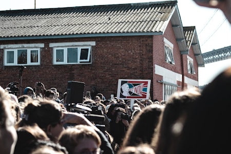A large crowd of people gathered outdoors, many holding cameras or their phones aloft, appears to be at some sort of public event or protest. Prominently displayed in the center is a sign with the words 'ACT NOW' against a stylized background featuring a profile image. In the background, there is a red brick building with white window frames and a corrugated roof, as well as a partial view of a bridge.