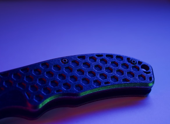 A close-up view of a knife handle with a hexagonal pattern, illuminated by blue and purple lighting. The surface texture and intricate design of the handle are highlighted, creating an interesting visual focus.