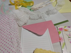 Various stationery items are laid out on a surface, including polka dot wrapping paper, a piece of white lace, a pink envelope, a sheet with floral design, and a yellow ribbon. There are also notepads, one with writing, and a card with a drawn bunny.