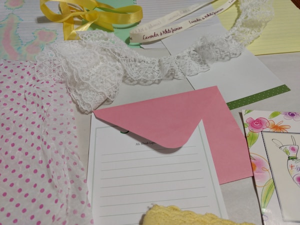 Various stationery items are laid out on a surface, including polka dot wrapping paper, a piece of white lace, a pink envelope, a sheet with floral design, and a yellow ribbon. There are also notepads, one with writing, and a card with a drawn bunny.