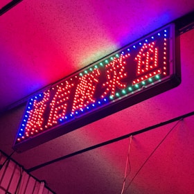 A vibrant, neon sign with red characters outlined by blue and green lights. It is mounted on a wall and the lighting creates a colorful glow on the surrounding area.