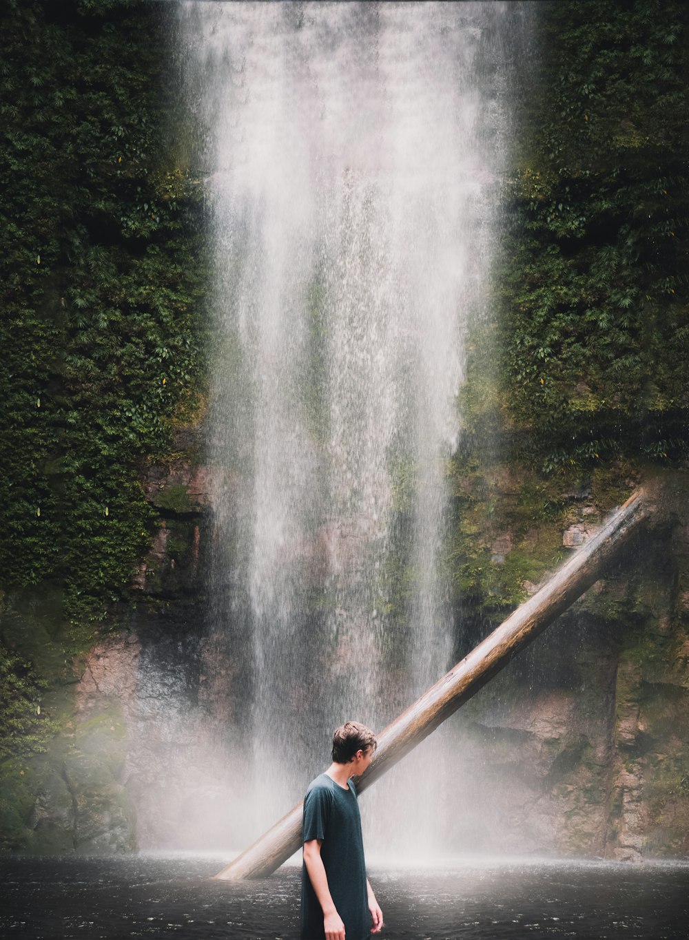 man in blue jacket and black pants standing on brown wooden bridge near waterfalls during daytime