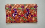 A rectangular fabric piece features intricate and colorful embroidery with floral and paisley patterns. The background is a rich orange shade, and the embroidery includes vibrant hues of purple, green, yellow, and pink with threads forming detailed and symmetrical designs.