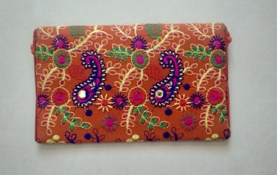 A rectangular fabric piece features intricate and colorful embroidery with floral and paisley patterns. The background is a rich orange shade, and the embroidery includes vibrant hues of purple, green, yellow, and pink with threads forming detailed and symmetrical designs.