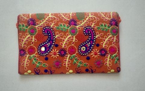 A rectangular fabric piece features intricate and colorful embroidery with floral and paisley patterns. The background is a rich orange shade, and the embroidery includes vibrant hues of purple, green, yellow, and pink with threads forming detailed and symmetrical designs.