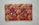 A rectangular fabric piece features intricate and colorful embroidery with floral and paisley patterns. The background is a rich orange shade, and the embroidery includes vibrant hues of purple, green, yellow, and pink with threads forming detailed and symmetrical designs.