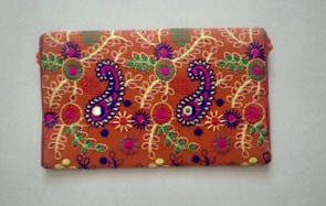 A rectangular fabric piece features intricate and colorful embroidery with floral and paisley patterns. The background is a rich orange shade, and the embroidery includes vibrant hues of purple, green, yellow, and pink with threads forming detailed and symmetrical designs.