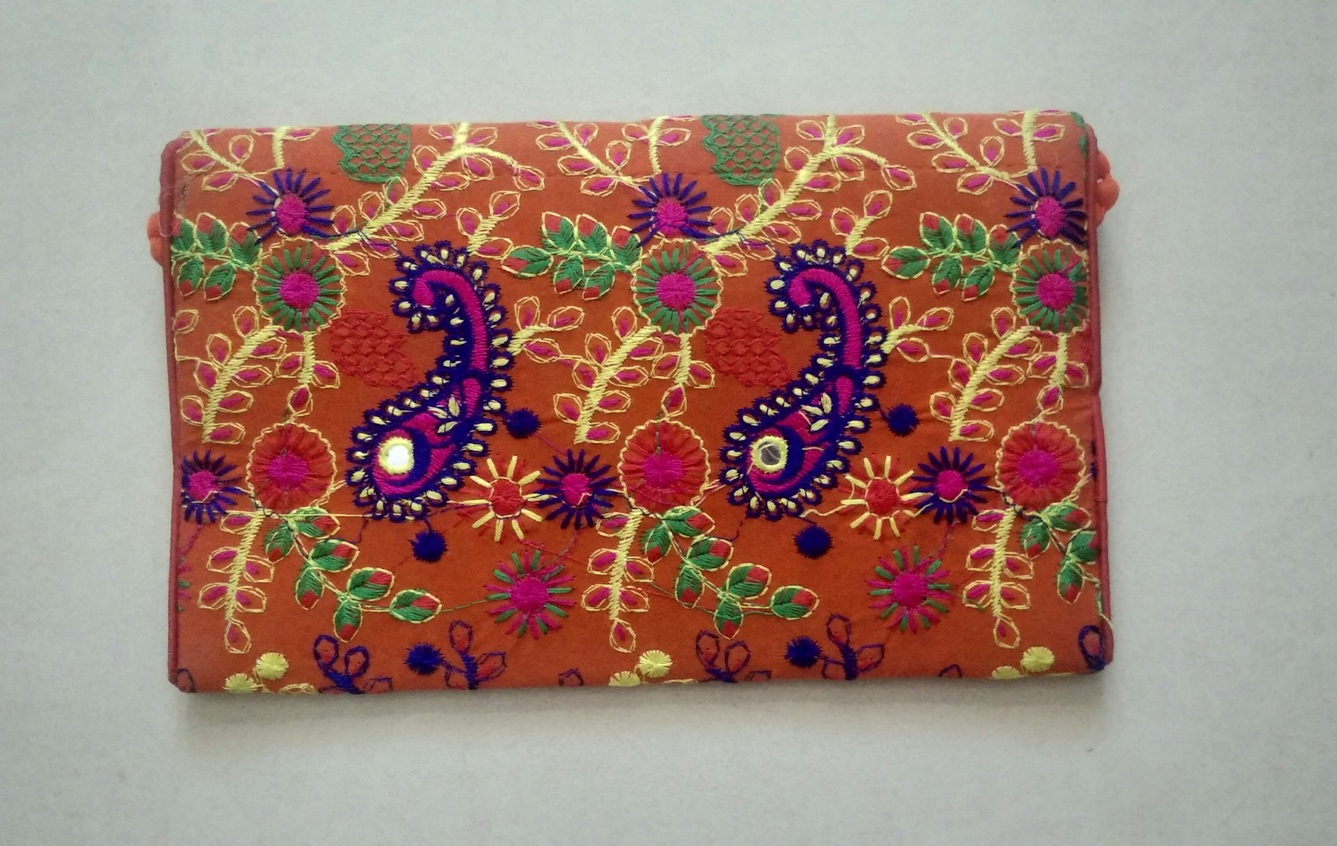 A rectangular fabric piece features intricate and colorful embroidery with floral and paisley patterns. The background is a rich orange shade, and the embroidery includes vibrant hues of purple, green, yellow, and pink with threads forming detailed and symmetrical designs.