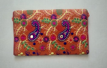 A rectangular fabric piece features intricate and colorful embroidery with floral and paisley patterns. The background is a rich orange shade, and the embroidery includes vibrant hues of purple, green, yellow, and pink with threads forming detailed and symmetrical designs.