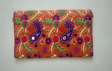 A rectangular fabric piece features intricate and colorful embroidery with floral and paisley patterns. The background is a rich orange shade, and the embroidery includes vibrant hues of purple, green, yellow, and pink with threads forming detailed and symmetrical designs.