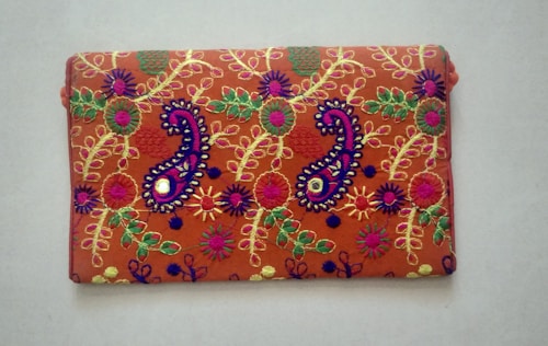 A rectangular fabric piece features intricate and colorful embroidery with floral and paisley patterns. The background is a rich orange shade, and the embroidery includes vibrant hues of purple, green, yellow, and pink with threads forming detailed and symmetrical designs.