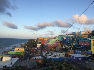 Colorful houses stretch along a coastal hillside, contrasting against the deep blue sea and sky. The vibrant buildings in shades of blue, pink, yellow, and green create a lively and picturesque scene. Palm trees are scattered throughout, adding a tropical vibe.