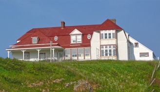 A large two-story house with a red roof and white exterior walls. The architectural design includes multiple windows, some with decorative grids, and a wrap-around porch supported by white columns. It is situated on a hill with green grass and patches of wildflowers in the foreground and a clear blue sky in the background.