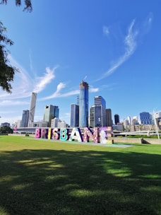 A vibrant cityscape with modern skyscrapers against a clear blue sky. Large colorful letters spelling 'BRISBANE' are prominently displayed on a green lawn in the foreground. The buildings include a mix of under-construction and completed high-rise structures.
