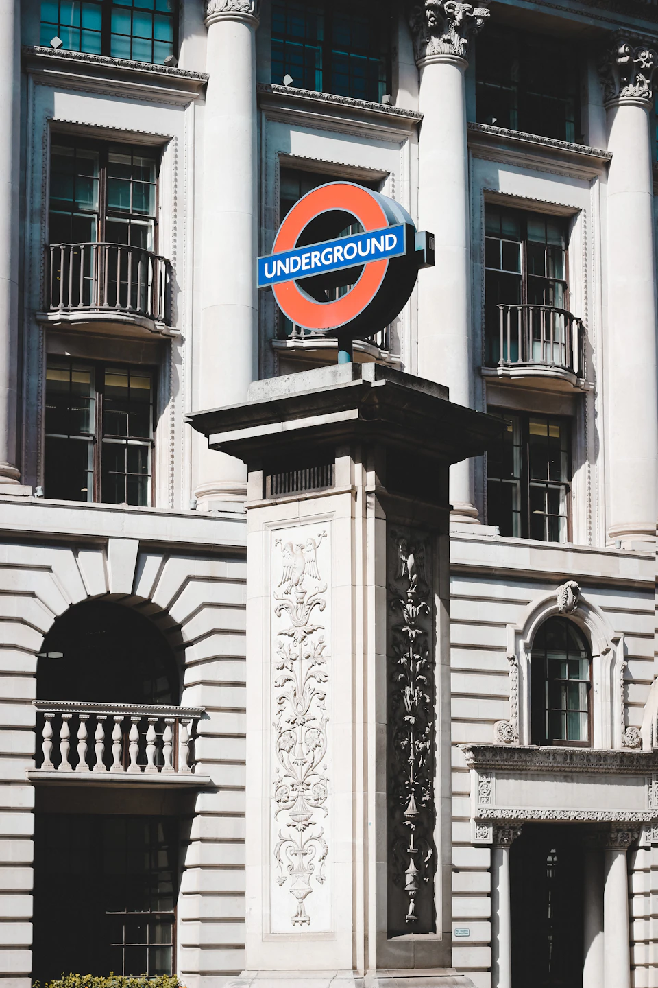 The image shows a tall, ornate white building with columns and arches, standing against a clear blue sky. The building features a large, circular sign on its side that reads "Underground" in bold red letters. A smaller sign below the main sign indicates the Underground station's location. The building is situated next to a sidewalk, suggesting it may be part of a larger complex or city center.