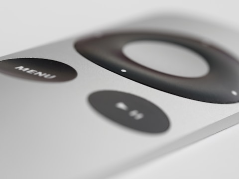A close-up view of a sleek, silver remote control with a prominent black circular navigation button and a 'MENU' button. The design is minimalist and modern.