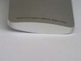 A close-up of a metallic device with smooth, rounded edges. The surface is silver-colored with a fine texture. Text printed on it reads: 'Designed by Apple in California Made in China.'