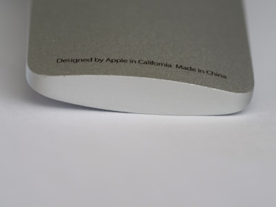 A close-up of a metallic device with smooth, rounded edges. The surface is silver-colored with a fine texture. Text printed on it reads: 'Designed by Apple in California Made in China.'