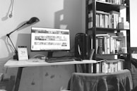 A black-and-white photo depicts a tidy home office setup. A computer monitor displays a webpage on a white desk, accompanied by a keyboard, mouse, and a desk lamp. A calendar sits to the left side of the desk. Behind the setup is a tall bookshelf filled with various books. A chair with a piece of clothing draped over it is visible in the foreground.