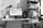 A black-and-white photo depicts a tidy home office setup. A computer monitor displays a webpage on a white desk, accompanied by a keyboard, mouse, and a desk lamp. A calendar sits to the left side of the desk. Behind the setup is a tall bookshelf filled with various books. A chair with a piece of clothing draped over it is visible in the foreground.