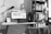 A black-and-white photo depicts a tidy home office setup. A computer monitor displays a webpage on a white desk, accompanied by a keyboard, mouse, and a desk lamp. A calendar sits to the left side of the desk. Behind the setup is a tall bookshelf filled with various books. A chair with a piece of clothing draped over it is visible in the foreground.