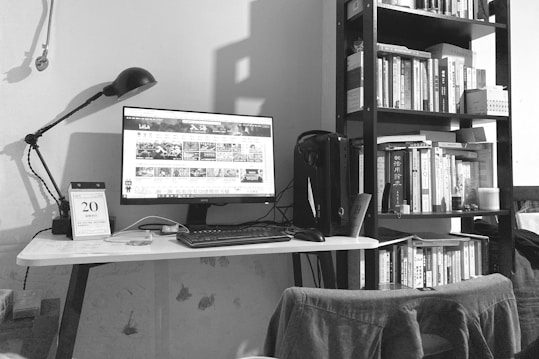 A black-and-white photo depicts a tidy home office setup. A computer monitor displays a webpage on a white desk, accompanied by a keyboard, mouse, and a desk lamp. A calendar sits to the left side of the desk. Behind the setup is a tall bookshelf filled with various books. A chair with a piece of clothing draped over it is visible in the foreground.