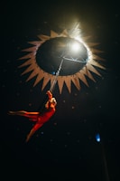 A performer in a bright red costume is suspended mid-air in a dark space, seemingly inside a circus tent. The background is adorned with star patterns and a circular light opening overhead, creating a dramatic spotlight effect on the performer.