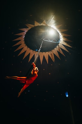 A performer in a bright red costume is suspended mid-air in a dark space, seemingly inside a circus tent. The background is adorned with star patterns and a circular light opening overhead, creating a dramatic spotlight effect on the performer.