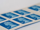 A strip of blue postage stamps featuring a profile image of a monarch, bordered by cream-colored edges with fine text. The stamps display '2nd' in the bottom corner, indicating a specific postage class.