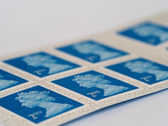 A strip of blue postage stamps featuring a profile image of a monarch, bordered by cream-colored edges with fine text. The stamps display '2nd' in the bottom corner, indicating a specific postage class.