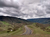 A panoramic view of a winding gravel road cutting through rolling hills under a cloudy sky.