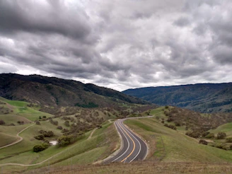 A panoramic view of a winding gravel road cutting through rolling hills under a cloudy sky.