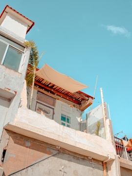 A partially constructed or renovated building with exposed walls and a canopy on the top floor. The structure shows unfinished areas and some construction materials. A few plants are visible, and a clear blue sky enhances the background.