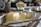 A block of butter partially cut into slices rests on an open wrapper on a wooden table. In the background, there are blurred objects, including a glass bowl likely containing more butter. A knife lies beside the butter.