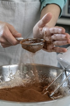 A person is sifting cocoa powder into a large metal bowl, where a mixer is also visible with batter partially mixed. The person&rsquo;s hands are partially covered in cocoa.