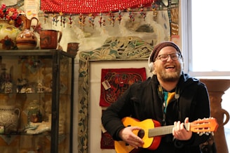 A man is smiling and playing a small acoustic guitar while wearing headphones. The scene is set in a vibrant room filled with eclectic decorations including pottery, textiles, and figurine ornaments. Warm tones of red, brown, and green are prominent in the decor, and there is a window beside him allowing light into the room.