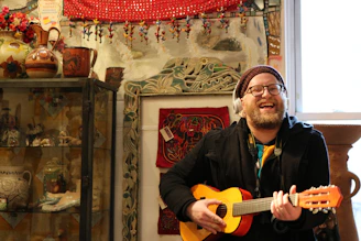 A smiling blind guitarist with short fingers playing a small hand guitar in a cozy, warmly lit room.
