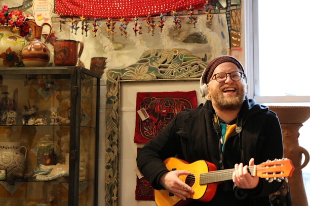 A man is smiling and playing a small acoustic guitar while wearing headphones. The scene is set in a vibrant room filled with eclectic decorations including pottery, textiles, and figurine ornaments. Warm tones of red, brown, and green are prominent in the decor, and there is a window beside him allowing light into the room.