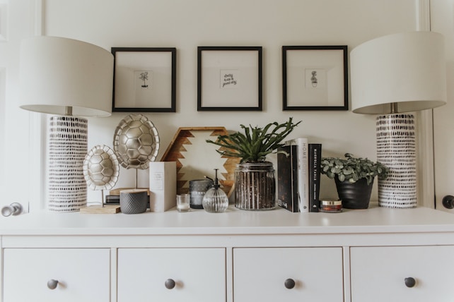 A chic dresser display featuring two modern lamps with patterned bases, several decorative ceramics with a textured design, framed minimalist art prints on the wall, various potted plants, and a selection of books and home décor items arranged neatly.
