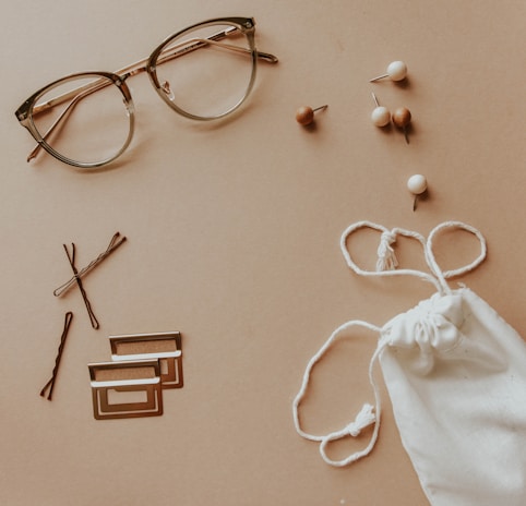 A pair of glasses is placed on a light tan surface alongside several small items. There are bobby pins, a few geometric paper clips, and thumbtacks with wooden and white spherical heads. On the right side, a white drawstring cloth pouch is positioned, with its strings casually arranged.