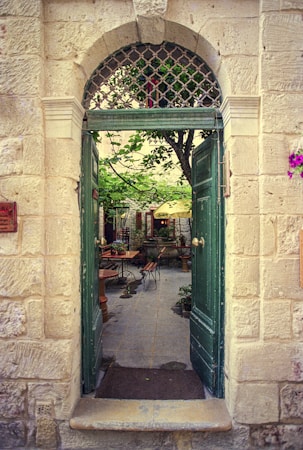 An open doorway reveals a charming patio with a quaint outdoor seating area. Green doors frame the view, and a tree in the center adds a natural touch. Several tables and chairs are arranged around the courtyard, with colorful flowers and plants enhancing the cozy atmosphere.