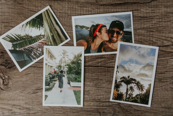 Four photographs are arranged on a wooden surface. One photo shows a close-up of palm leaves, another features a couple smiling with one person kissing the other on the cheek, a third depicts a woman walking with a child along a path lined with tropical plants, and the fourth captures a scenic view of palm trees against a partly cloudy sky.