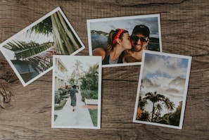 Four photographs are arranged on a wooden surface. One photo shows a close-up of palm leaves, another features a couple smiling with one person kissing the other on the cheek, a third depicts a woman walking with a child along a path lined with tropical plants, and the fourth captures a scenic view of palm trees against a partly cloudy sky.