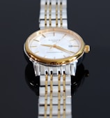 A close-up view of a luxury wristwatch featuring a gold and silver metal band. The watch has a round face with a white dial, golden hour markers, and hands. The brand name and model 'Tissot 1853 Powermatic 80' are visible on the dial.