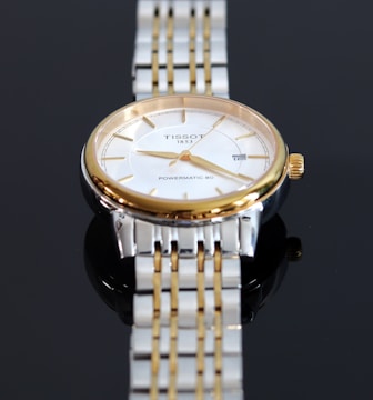 A close-up view of a luxury wristwatch featuring a gold and silver metal band. The watch has a round face with a white dial, golden hour markers, and hands. The brand name and model 'Tissot 1853 Powermatic 80' are visible on the dial.