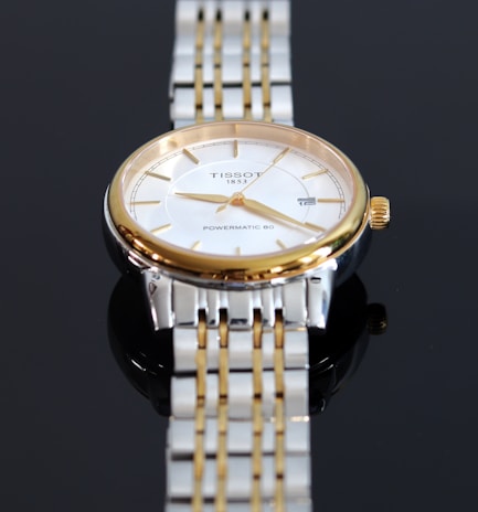 A close-up view of a luxury wristwatch featuring a gold and silver metal band. The watch has a round face with a white dial, golden hour markers, and hands. The brand name and model 'Tissot 1853 Powermatic 80' are visible on the dial.