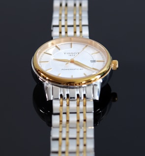 A close-up view of a luxury wristwatch featuring a gold and silver metal band. The watch has a round face with a white dial, golden hour markers, and hands. The brand name and model 'Tissot 1853 Powermatic 80' are visible on the dial.