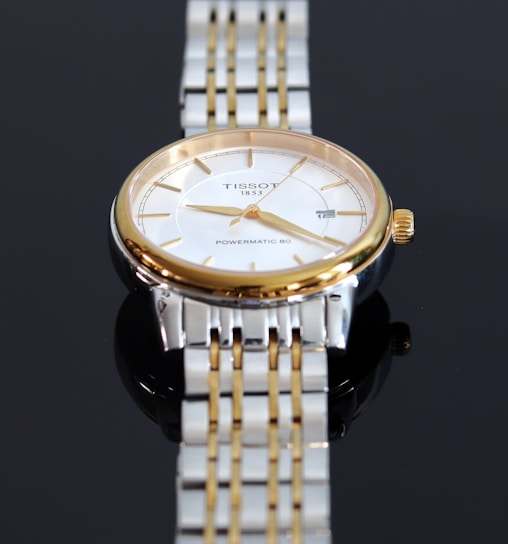 A close-up view of a luxury wristwatch featuring a gold and silver metal band. The watch has a round face with a white dial, golden hour markers, and hands. The brand name and model 'Tissot 1853 Powermatic 80' are visible on the dial.