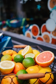 A colorful assortment of citrus fruits including lemons, limes, oranges, and grapefruits displayed in a wire basket. The background features a decorative window with pictures of sliced citrus fruits, creating a vibrant and fresh setting.